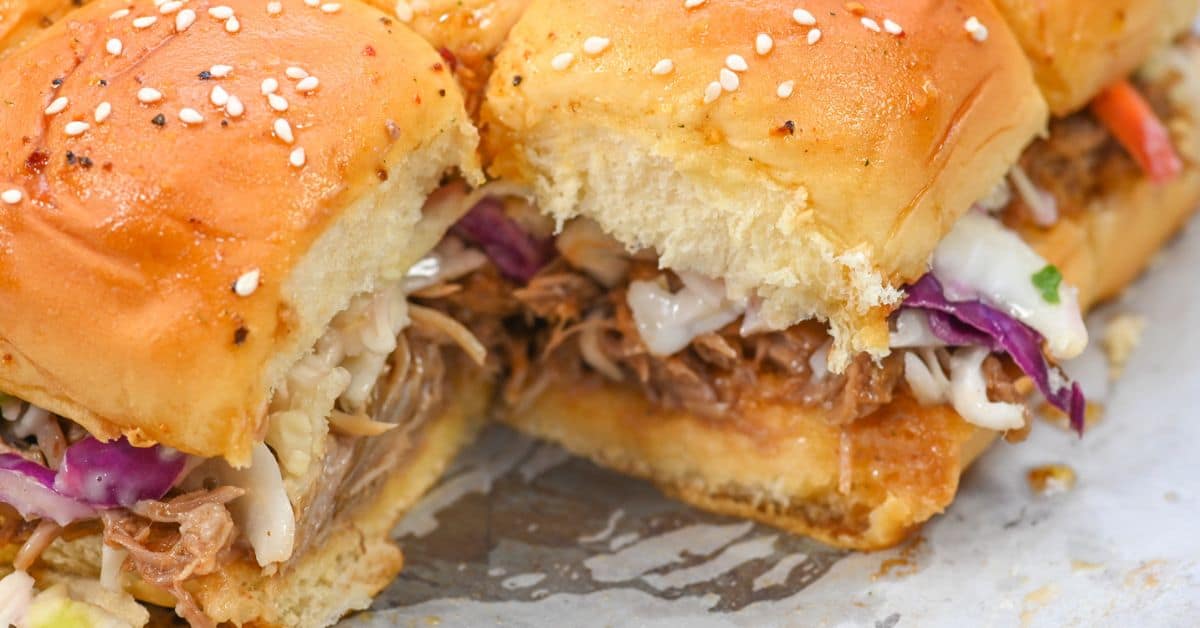 Close-up of pork sliders with coleslaw on sesame buns, featuring tender shredded pork, placed on a white surface.