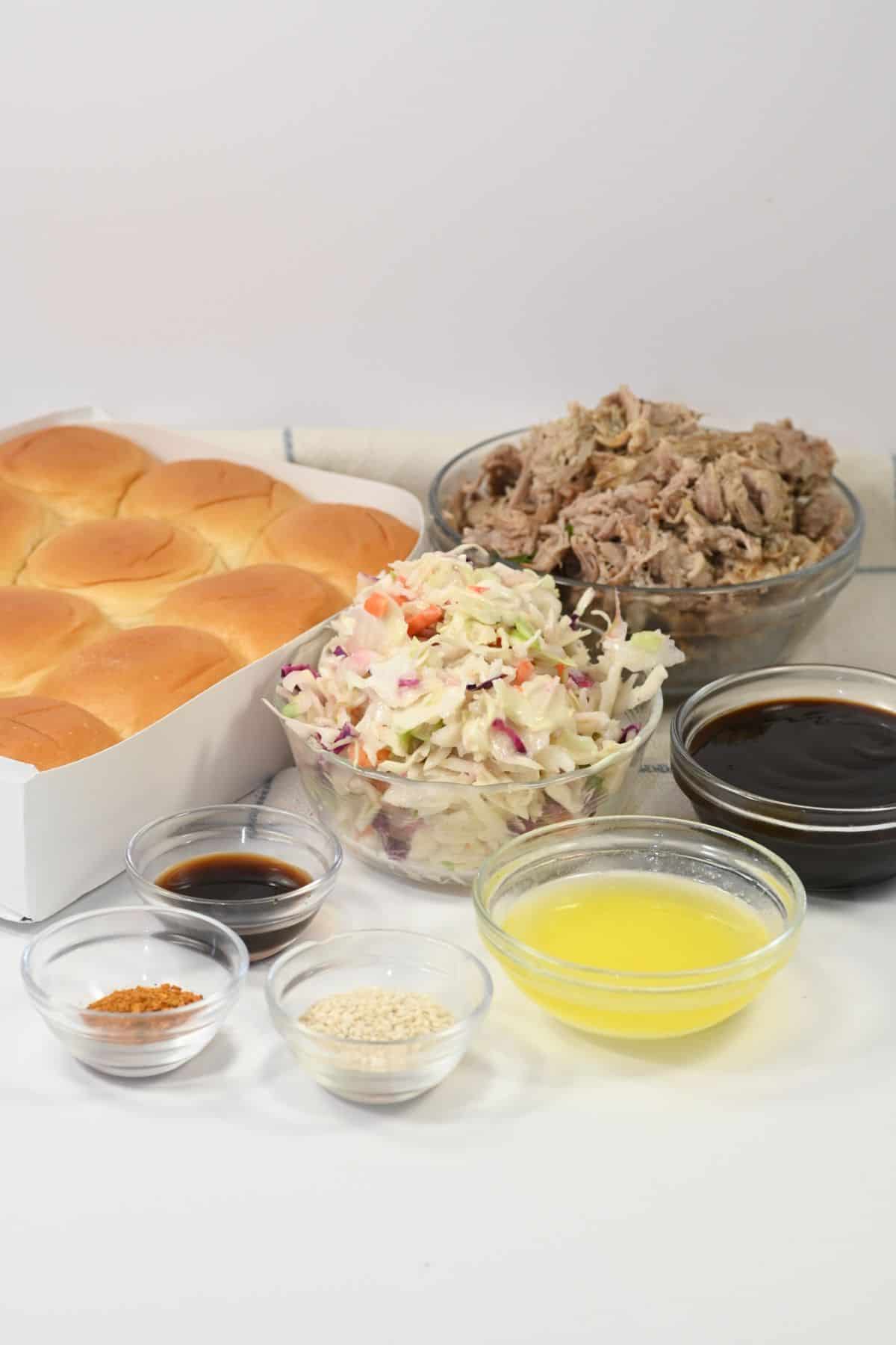 A tray of golden slider buns is paired with bowls of savory shredded pork, creamy coleslaw, barbecue sauce, melted butter, spices, sesame seeds, and soy sauce—all artfully arranged on a white surface to create the ultimate Pork Sliders experience.