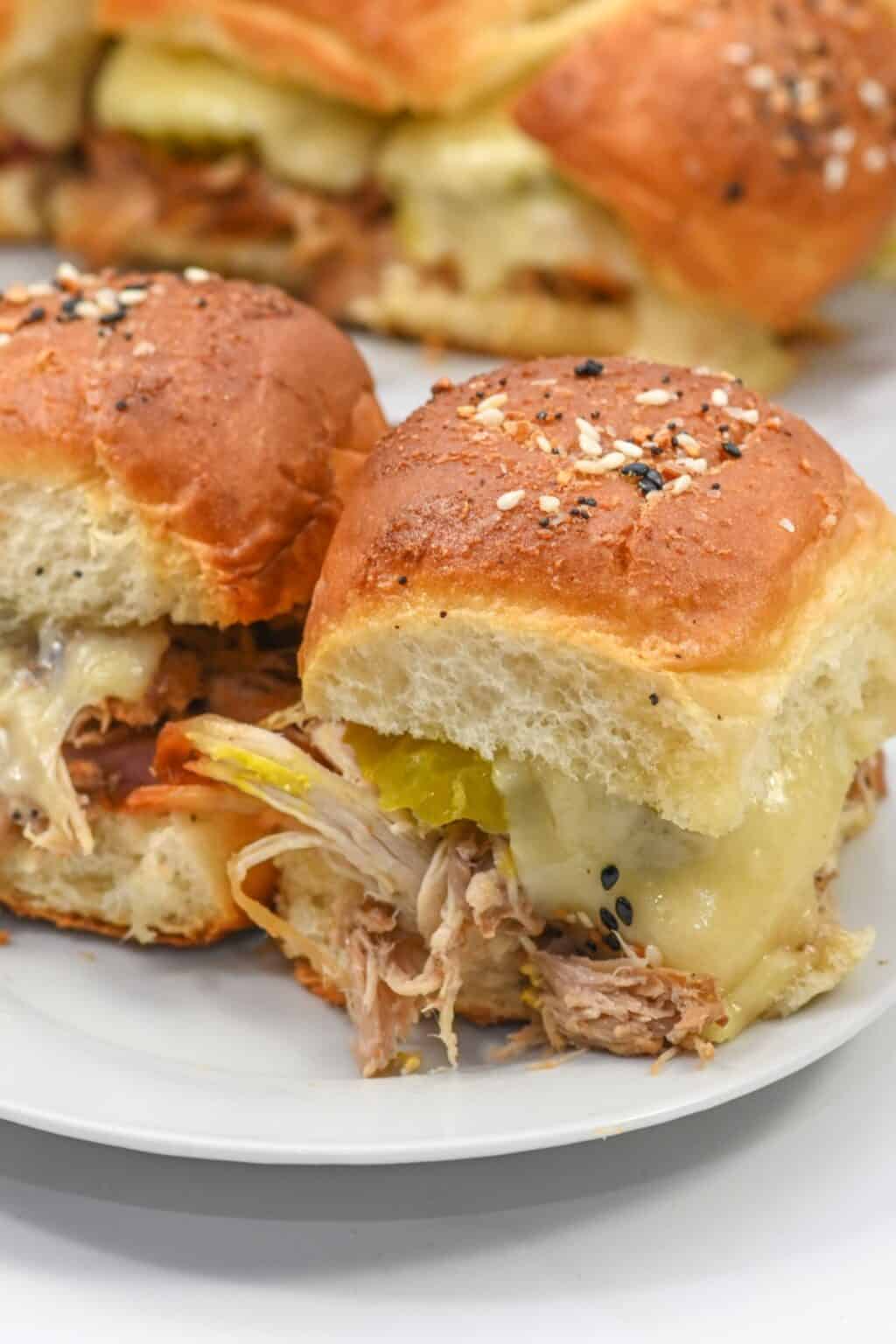 Two Cuban Sliders with pulled pork, melted cheese, and pickles on seeded buns, placed on a white plate with more sliders in the background.