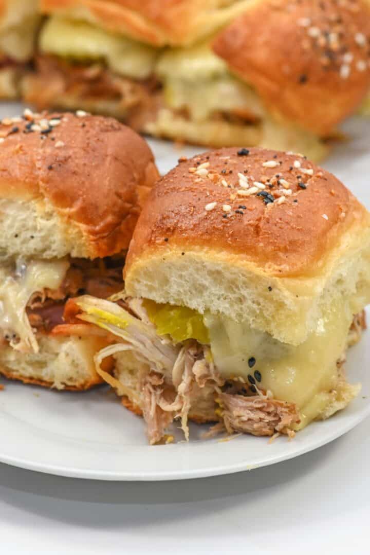 Two Cuban Sliders with pulled pork, melted cheese, and pickles on seeded buns, placed on a white plate with more sliders in the background.