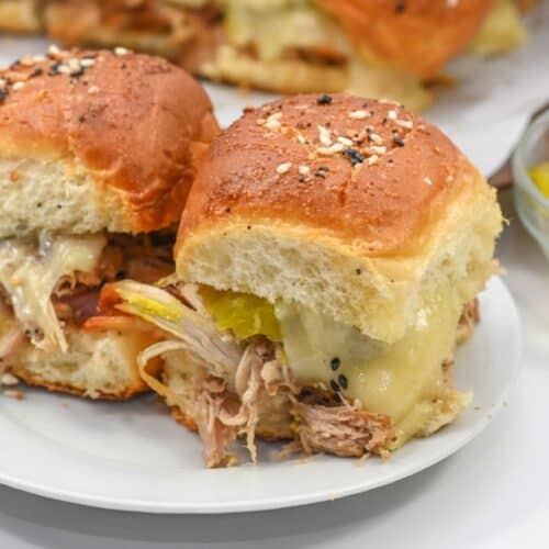 Two Cuban sliders piled high with tender pulled pork, melted cheese, and tangy pickle slices on a white plate, served alongside a small glass bowl of sliced pickles.