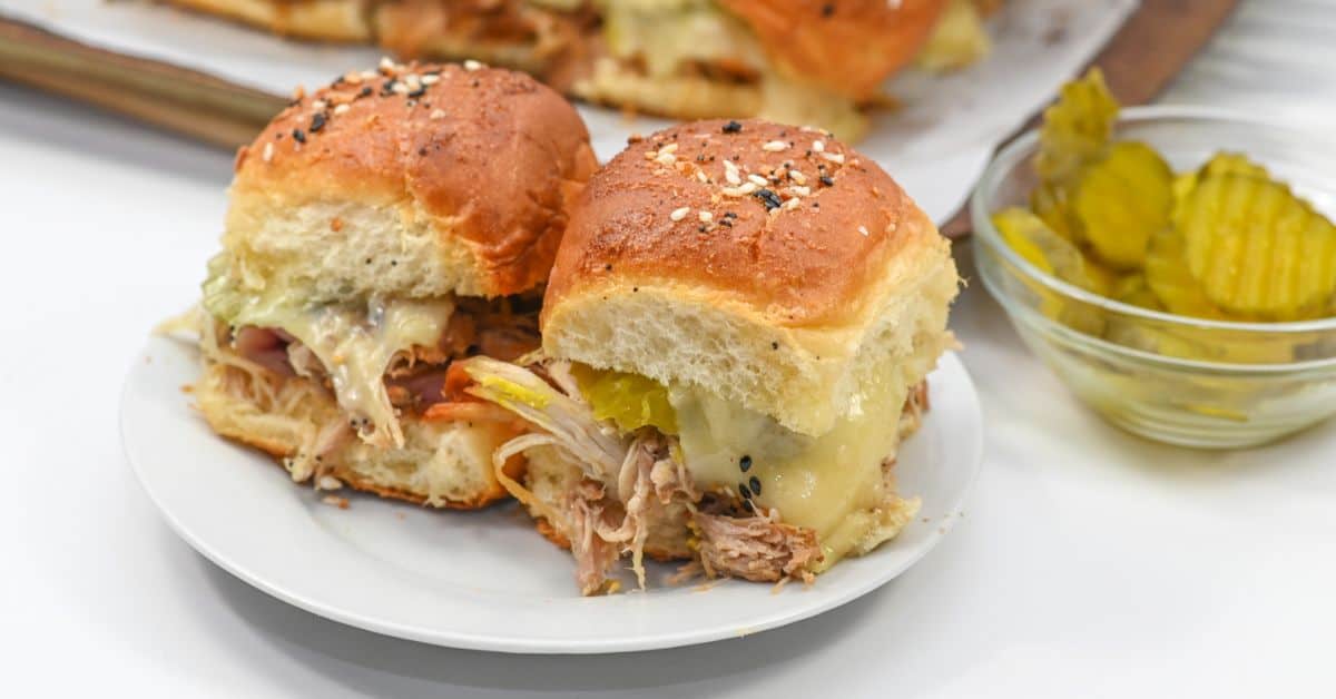 Two Cuban sliders piled high with tender pulled pork, melted cheese, and tangy pickle slices on a white plate, served alongside a small glass bowl of sliced pickles.