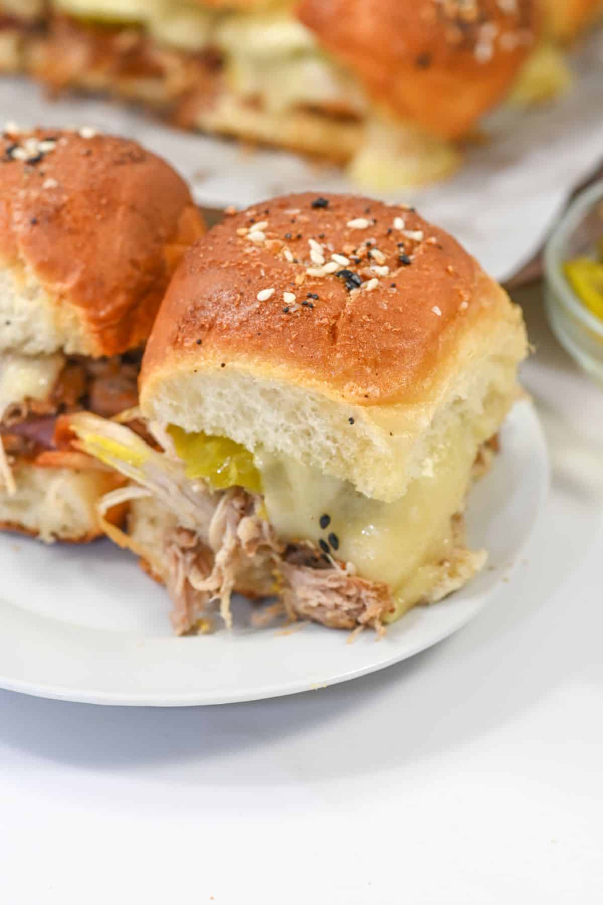 A Cuban Slider-inspired pulled pork slider with melted cheese and pickles on a seeded bun sits on a white plate, with more sliders and a small bowl of pickles in the background.