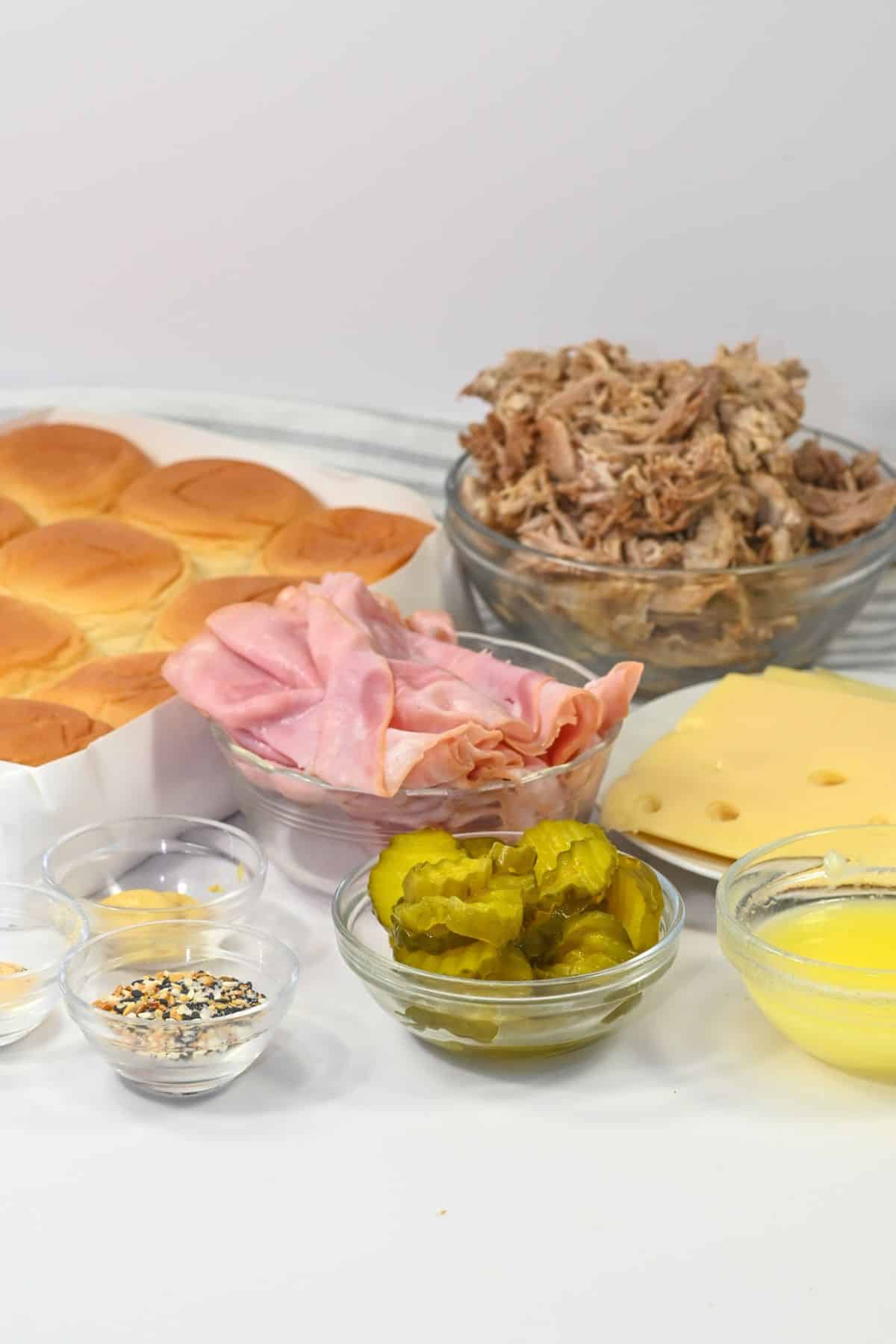 Ingredients for Cuban Sliders are arranged on a counter: slider buns, pulled pork, ham, Swiss cheese, pickles, melted butter, and seasonings in small bowls.
