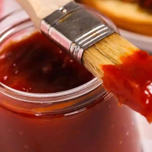 A close-up of a glass jar filled with homemade BBQ sauce, with a basting brush coated in the best BBQ sauce resting on the rim.