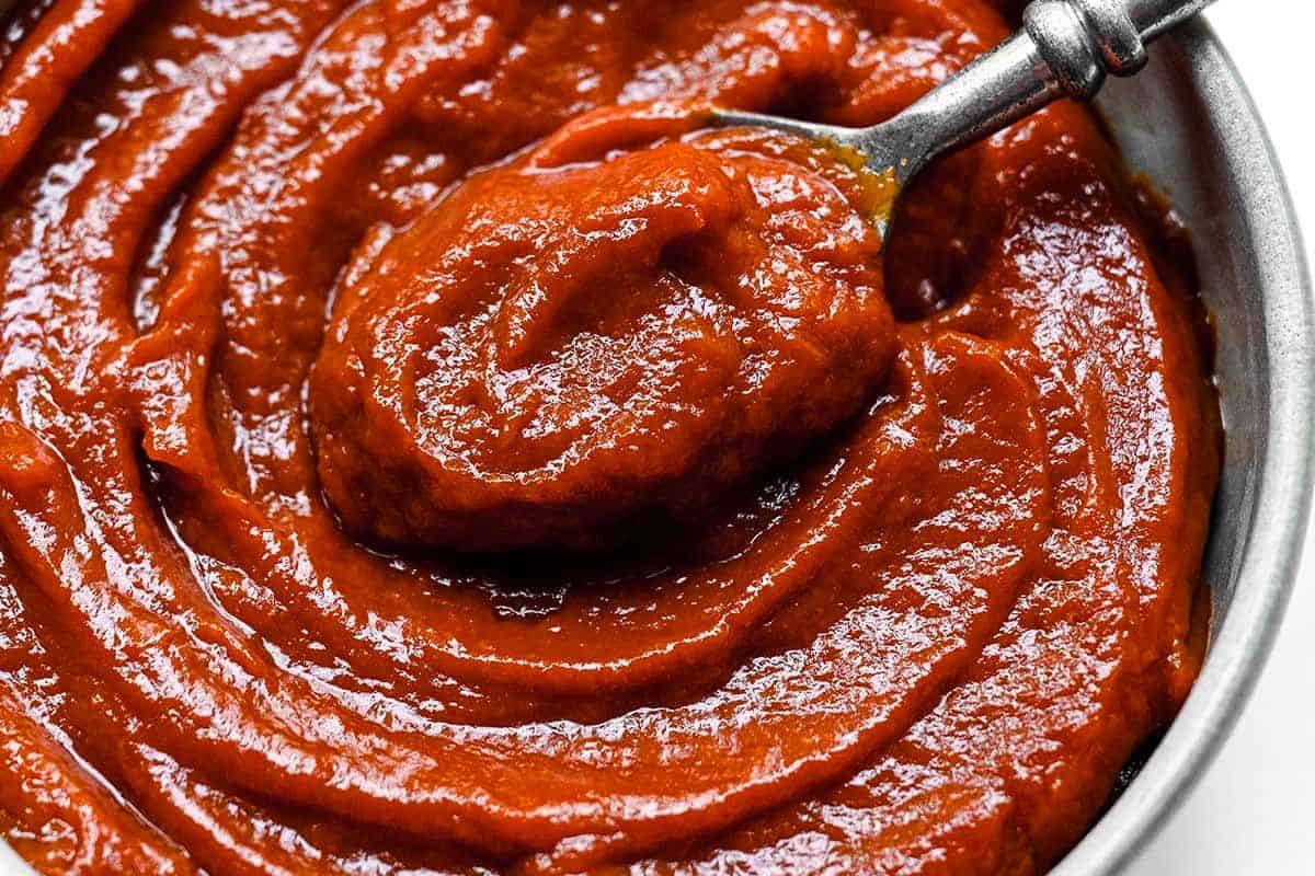 A close-up of a metal spoon scooping thick, red homemade BBQ sauce from a bowl.