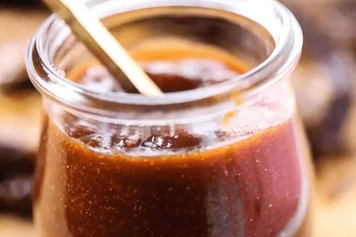 A close-up of a glass jar filled with thick, reddish-brown homemade BBQ sauce, with a spoon inside the jar.