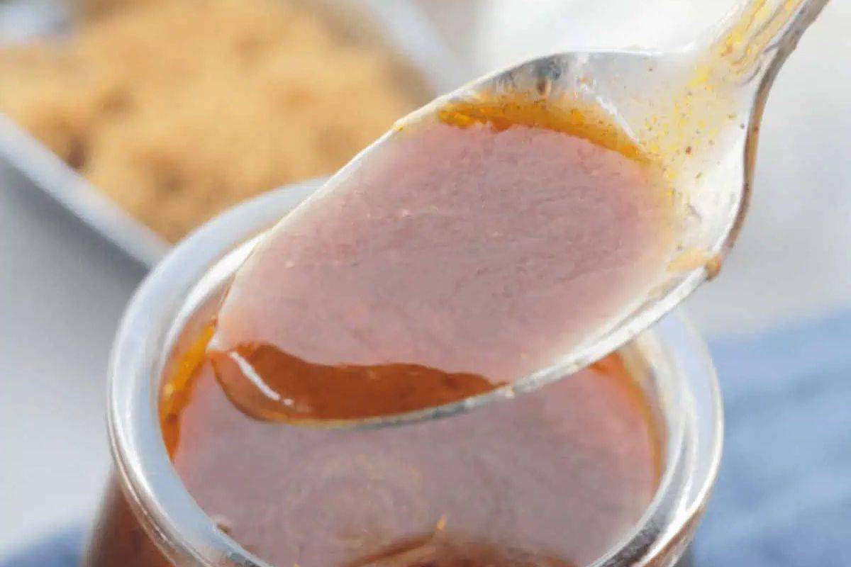 A spoon scoops thick brown barbecue sauce from a glass jar, with a blurred container of brown sugar in the background.