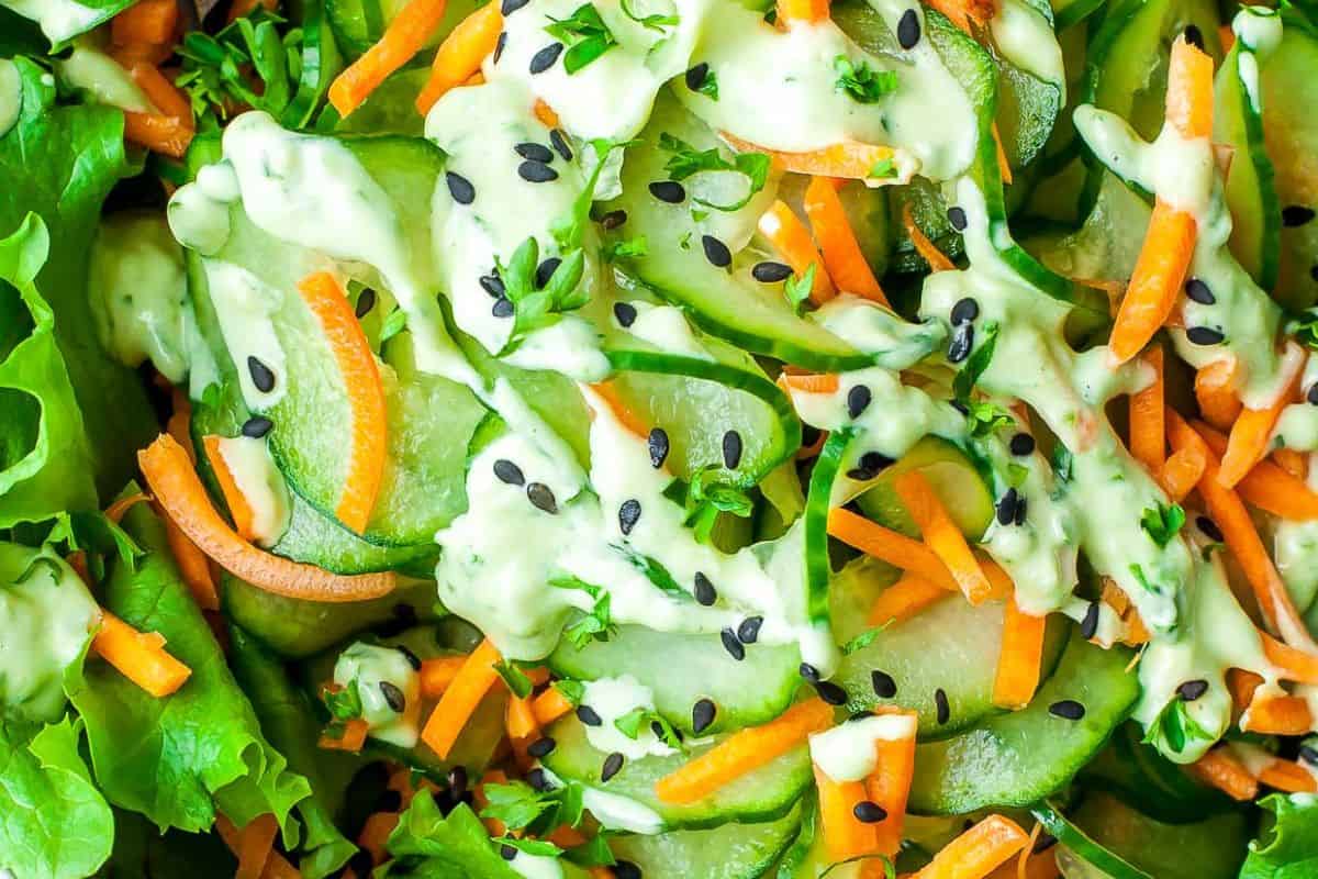 A close-up of a fresh salad with sliced cucumbers, shredded carrots, green lettuce, and black sesame seeds, all generously drizzled with a creamy, light green sauce, likely wasabi dressing.