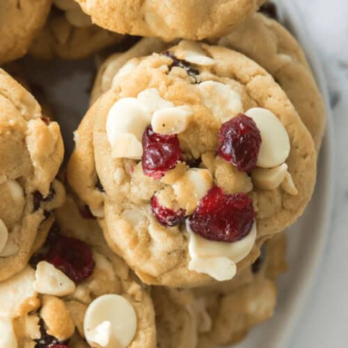 Close-up image of white chocolate cranberry cookies, featuring white chocolate chips and dried cranberries, stacked on a white plate.