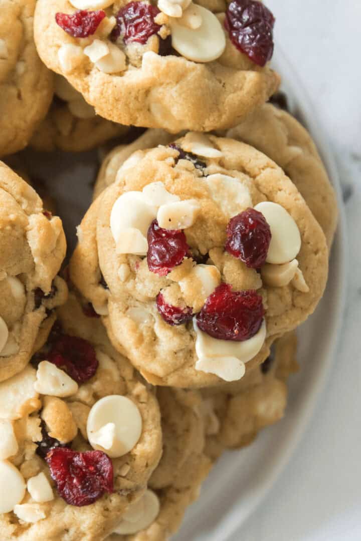Close-up image of white chocolate cranberry cookies, featuring white chocolate chips and dried cranberries, stacked on a white plate.
