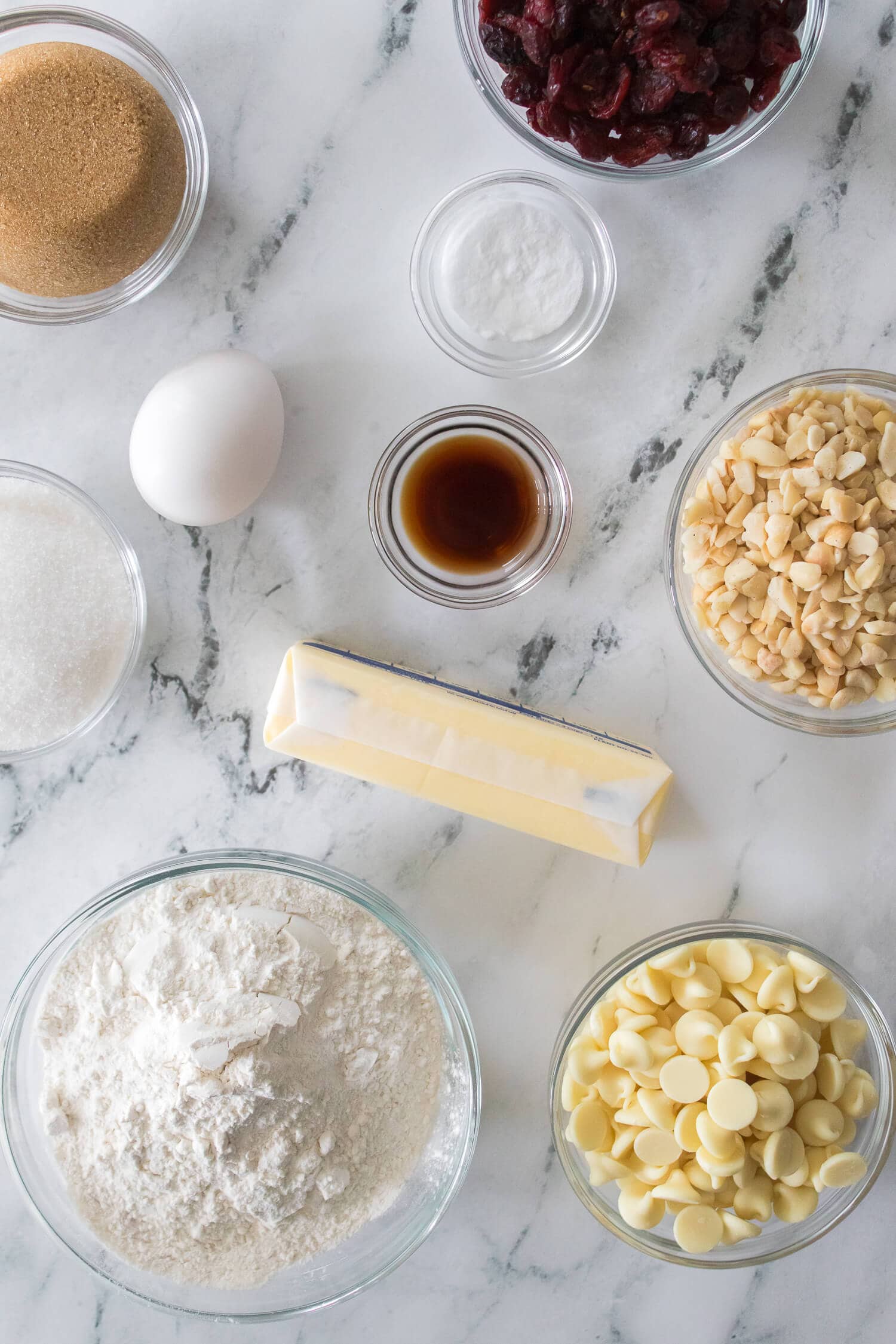 Ingredients for baking white chocolate cranberry cookies arranged on a marble surface include flour, white chocolate chips, chopped nuts, vanilla extract, brown sugar, granulated sugar, an egg, dried cranberries, baking soda, and a stick of butter.