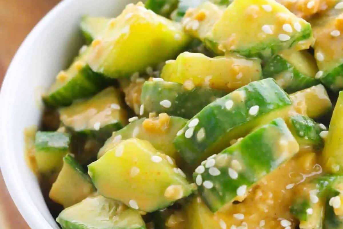 A close-up shot of a bowl of chunky cucumber slices coated in a creamy, yellowish-brown sauce and sprinkled generously with white sesame seeds.