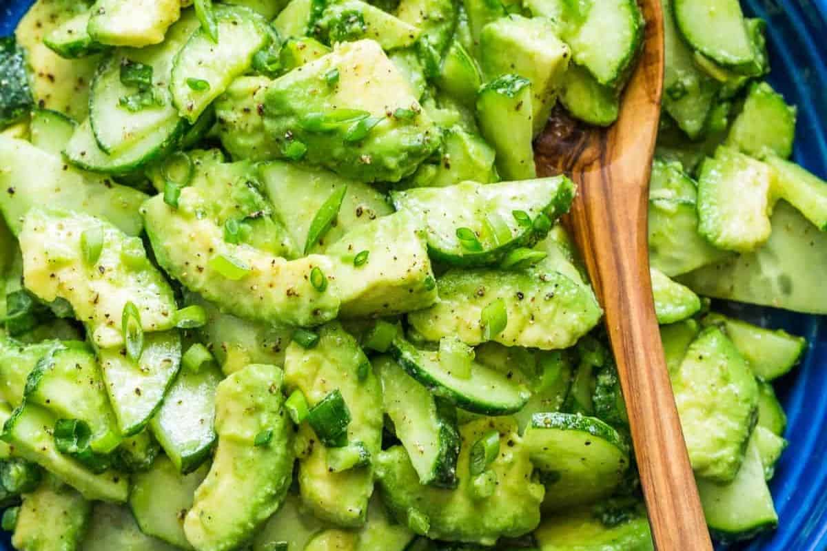 A bright green salad in a blue bowl with a wooden spoon, featuring chunks of avocado and sliced cucumbers, garnished with chopped green onions and a sprinkle of black pepper.