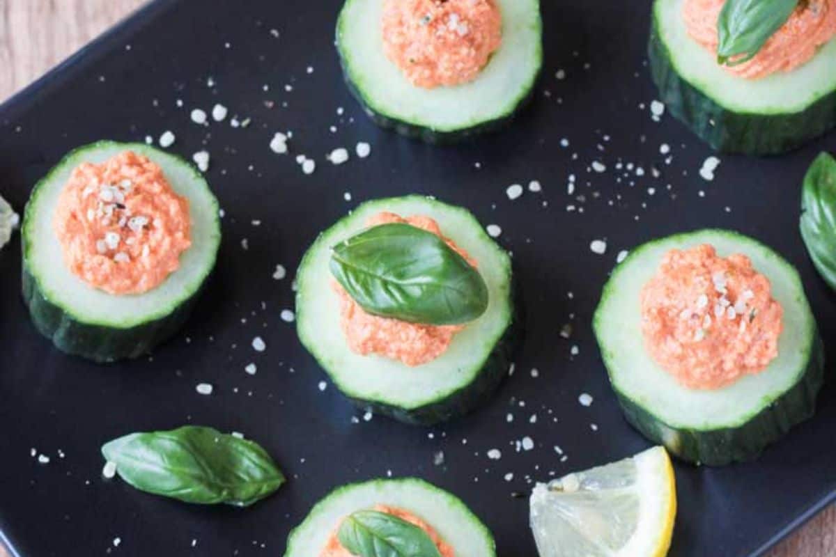 Several round cucumber slices topped with a vibrant orange, creamy mixture and garnished with fresh basil leaves and a sprinkle of salt, arranged on a dark plate with a lemon wedge.