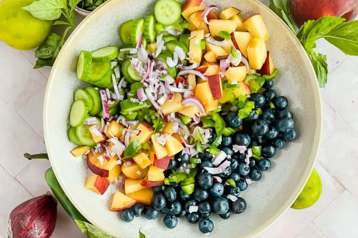 A large bowl filled with various chopped ingredients for a salad, including sliced cucumbers, diced red onions, chopped peaches, green bell peppers, and a mound of fresh blueberries, with basil leaves and limes around the bowl.