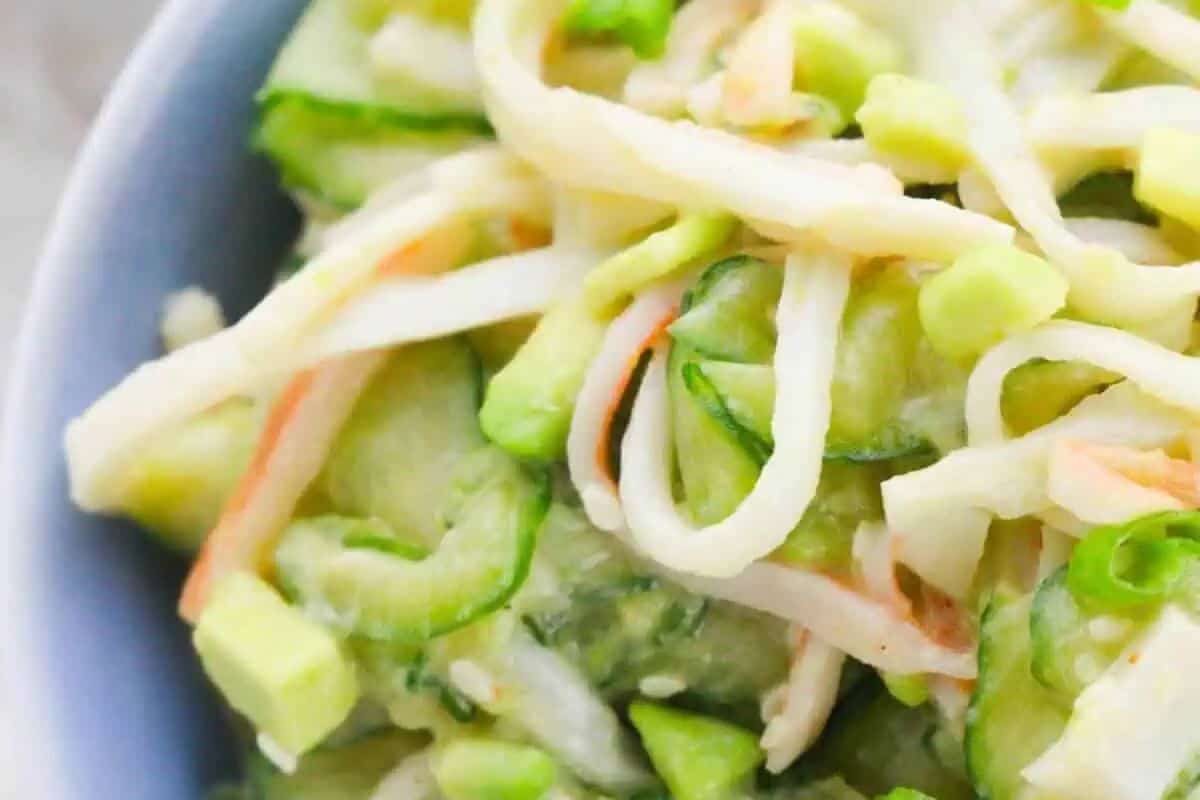  A close-up of a salad in a light blue bowl, featuring julienned cucumber, imitation crab sticks, and possibly avocado, all mixed in a light dressing.