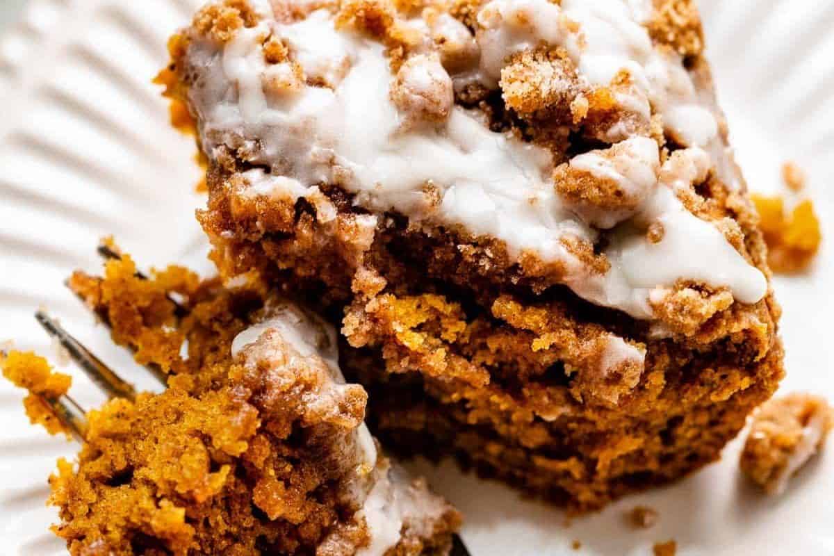 Close-up of a piece of crumbly cake with white icing on top, served on a white plate with a fork—a tempting treat for fans of irresistible recipes and pumpkin spice flavors.