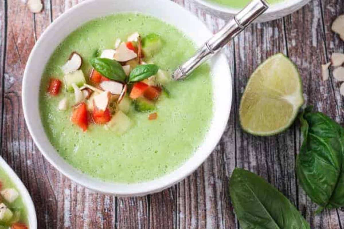 A white bowl of light green, creamy cold soup garnished with diced red tomatoes, chopped green cucumbers, sliced almonds, and fresh basil leaves, with a metal spoon, lime halves, and basil leaves on a rustic wooden table.