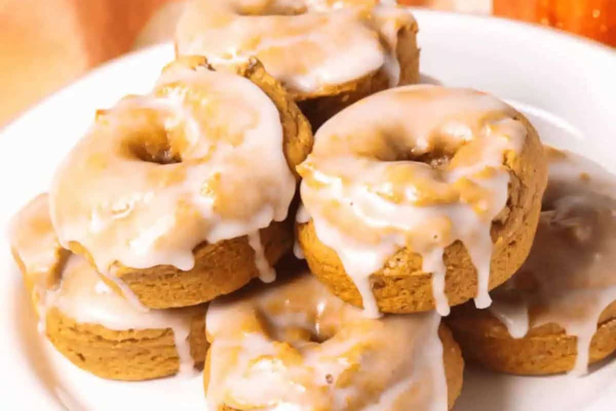 A plate stacked with pumpkin spice donuts, each topped with a layer of white icing that drips down the sides—a delicious addition to your favorite pumpkin recipes.