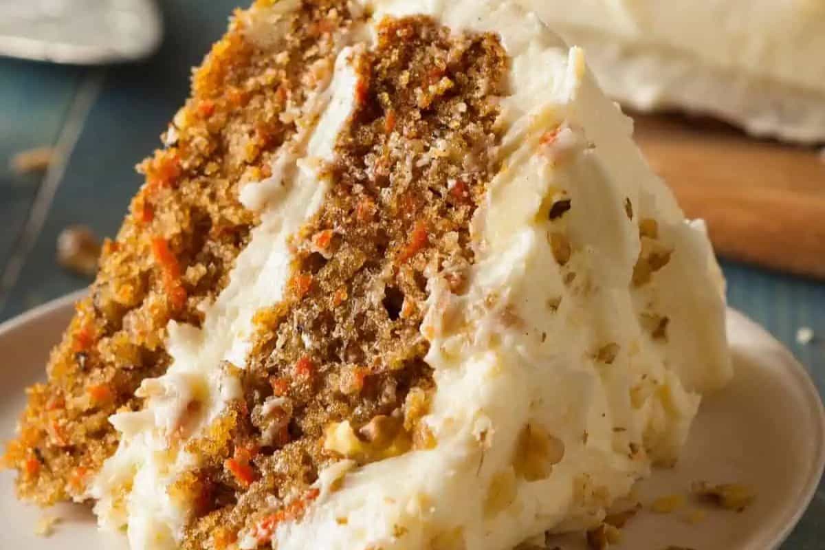 A slice of pumpkin carrot cake with cream cheese frosting and visible pieces of walnut sits on a white plate, perfect for fans of pumpkin spice recipes.