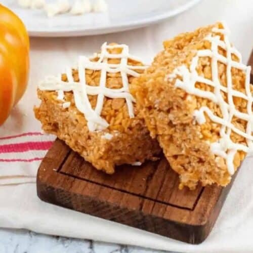 Two pumpkin rice krispie treats with white icing drizzled in a crisscross pattern sit on a small wooden board next to a decorative pumpkin, making them perfect for fans of irresistible pumpkin spice recipes.