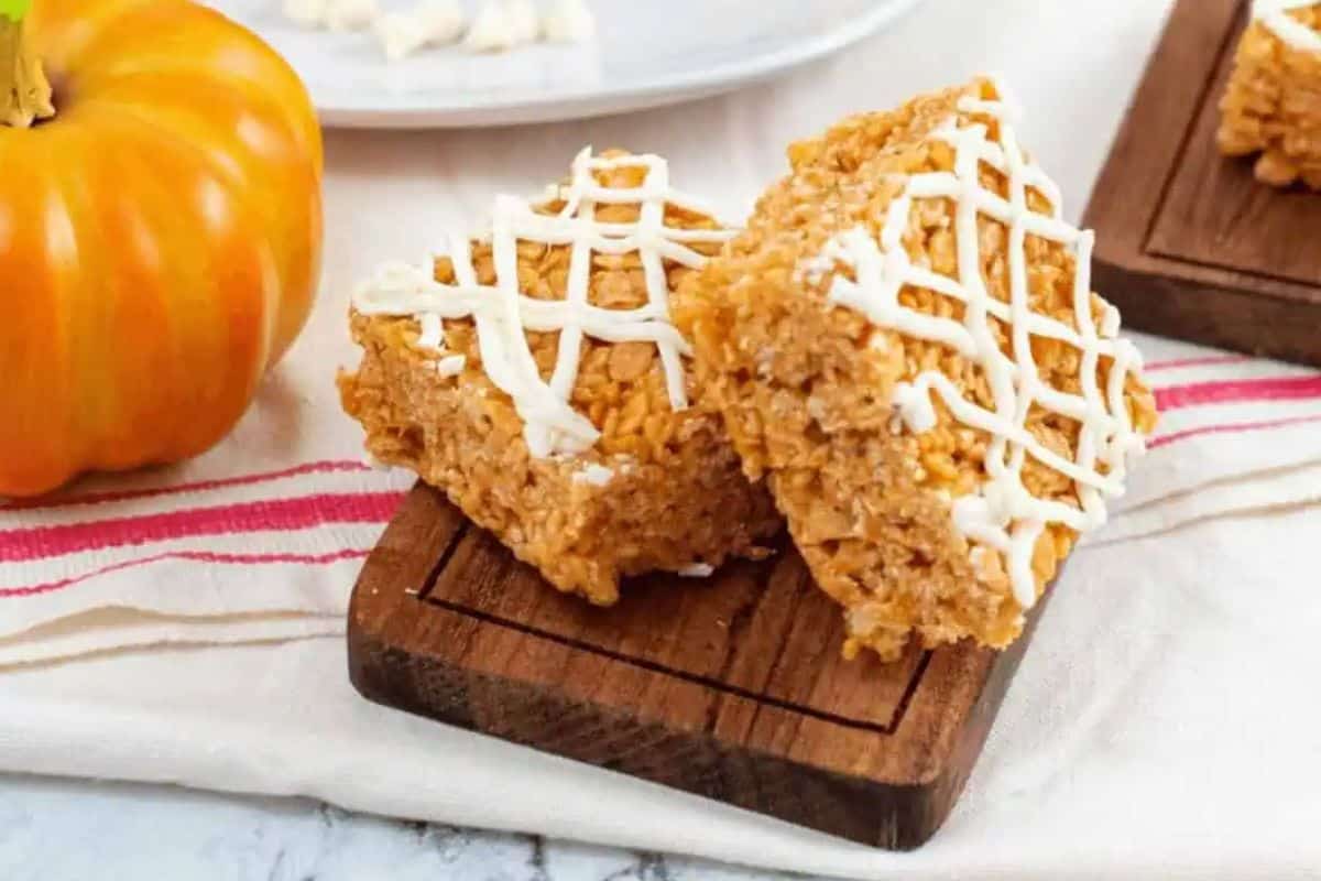 Two pumpkin rice krispie treats with white icing drizzled in a crisscross pattern sit on a small wooden board next to a decorative pumpkin, making them perfect for fans of irresistible pumpkin spice recipes.