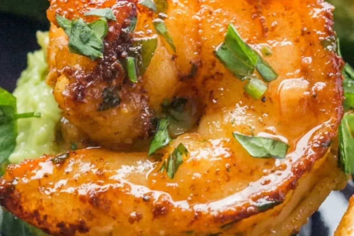 A close-up of several large, cooked shrimp with a golden-brown, slightly charred appearance, garnished with chopped green herbs, resting on a green, possibly avocado, base.
