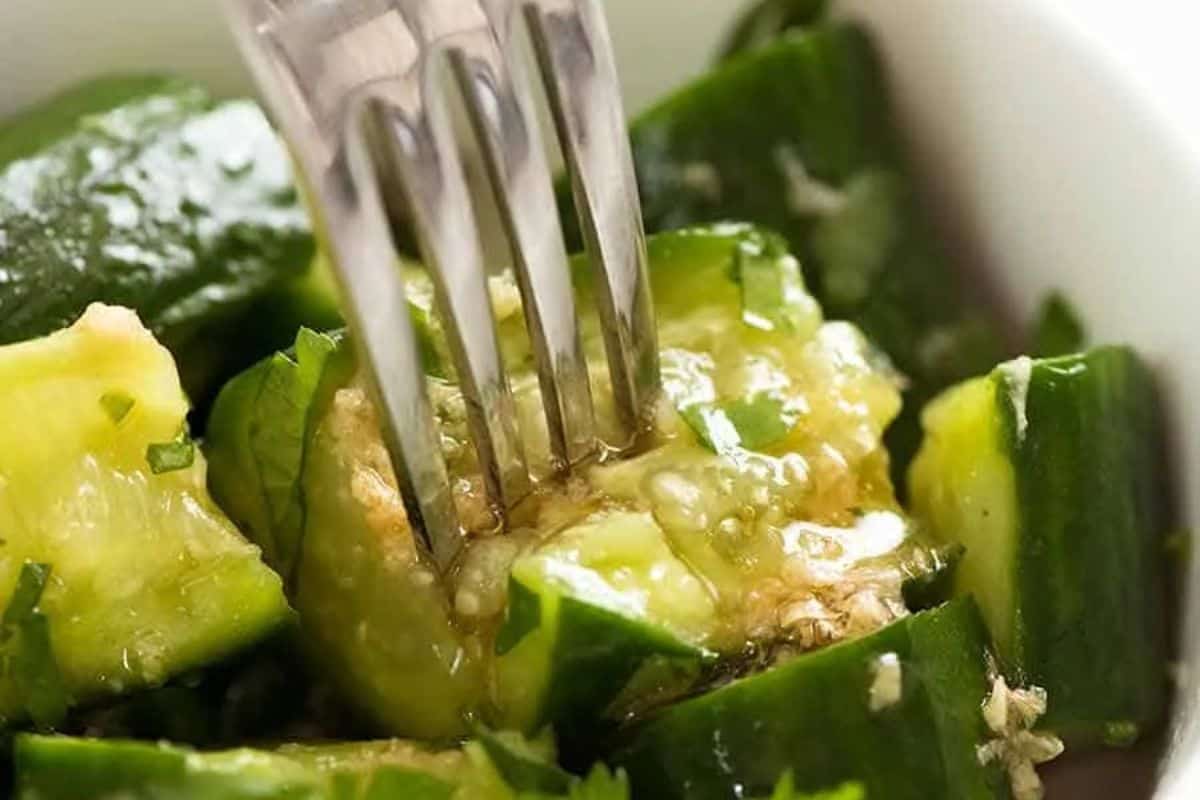  A fork piercing a piece of smashed cucumber in a bowl of what appears to be a cucumber salad with a light dressing and some herbs.