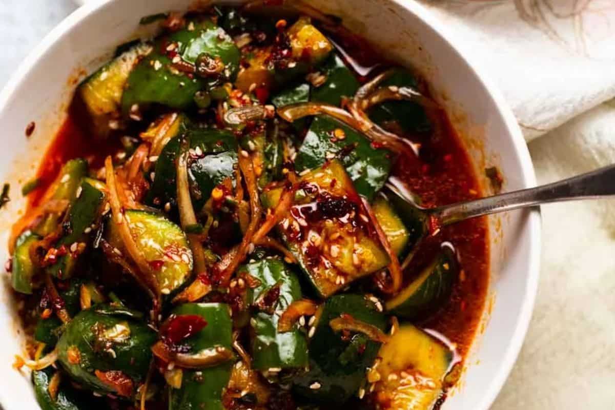 A white bowl filled with chunky cucumber pieces in a vibrant red, oily, spicy sauce, garnished with sesame seeds, and containing a spoon.