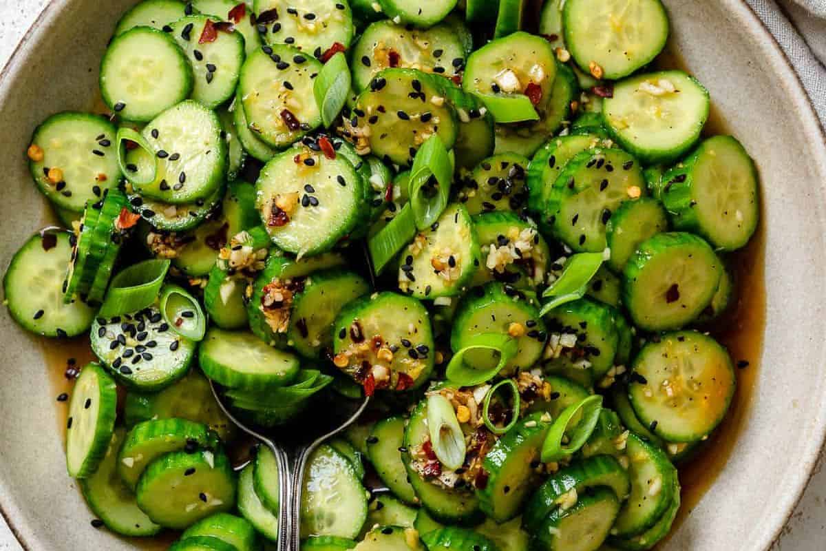 A bowl of thinly sliced cucumbers mixed with green onions, red pepper flakes, and black and white sesame seeds, in a light sauce, with a spoon resting in the bowl.
