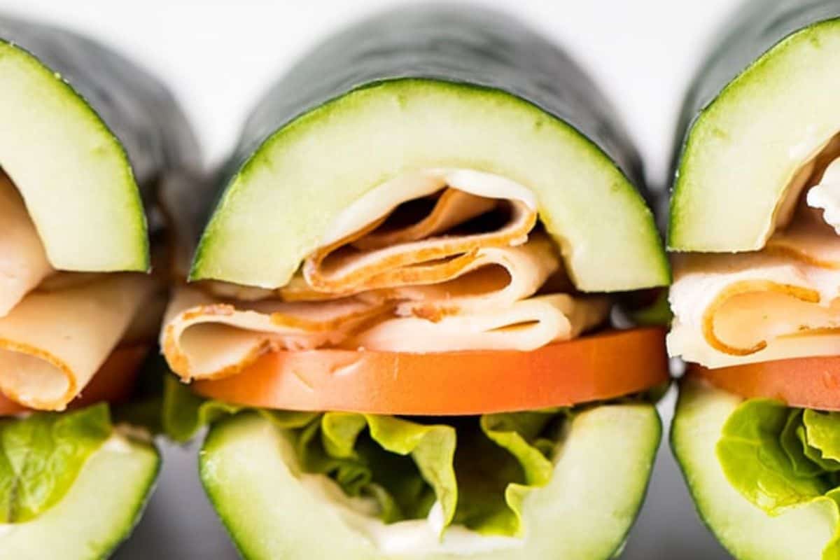 A close-up of three "subs" made from hollowed-out cucumbers serving as bread, filled with deli meat (possibly turkey), a slice of tomato, and green lettuce.