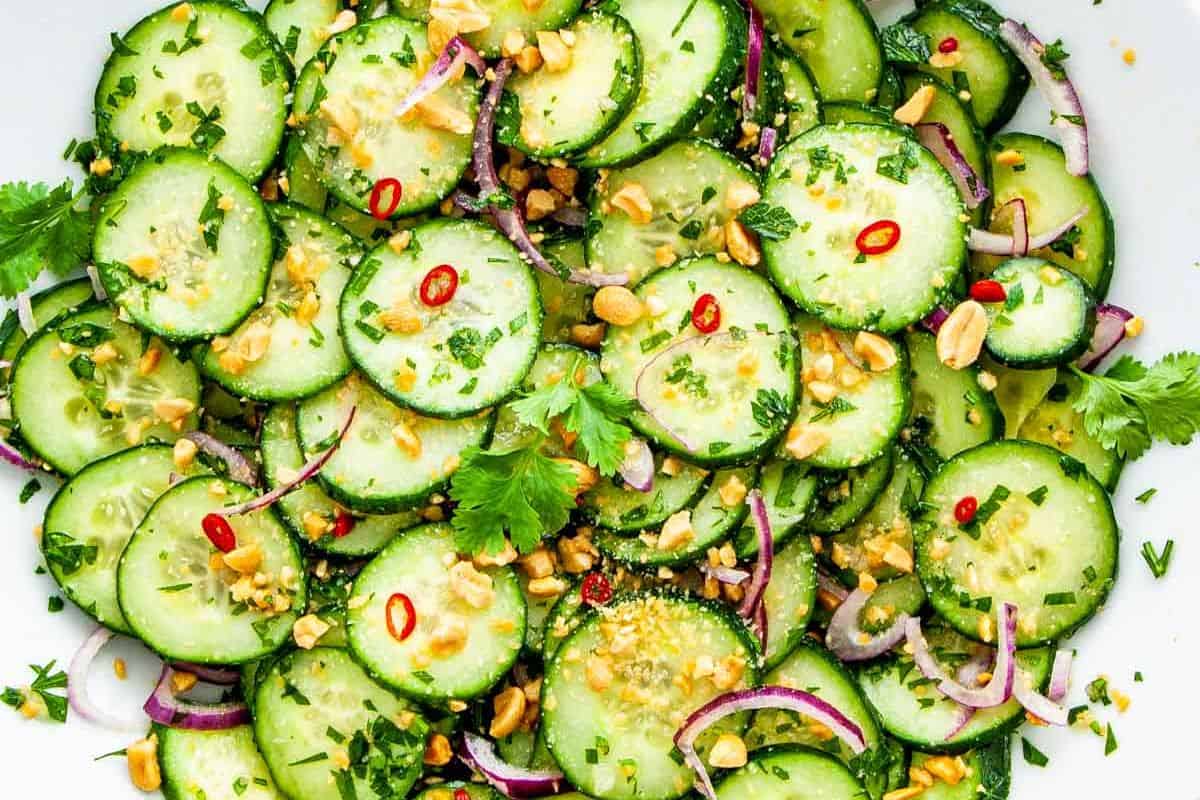 An overhead shot of a Thai-style cucumber salad with thinly sliced cucumbers, red onion rings, chopped peanuts, fresh herbs (cilantro and mint), and red chili slices, all mixed together.