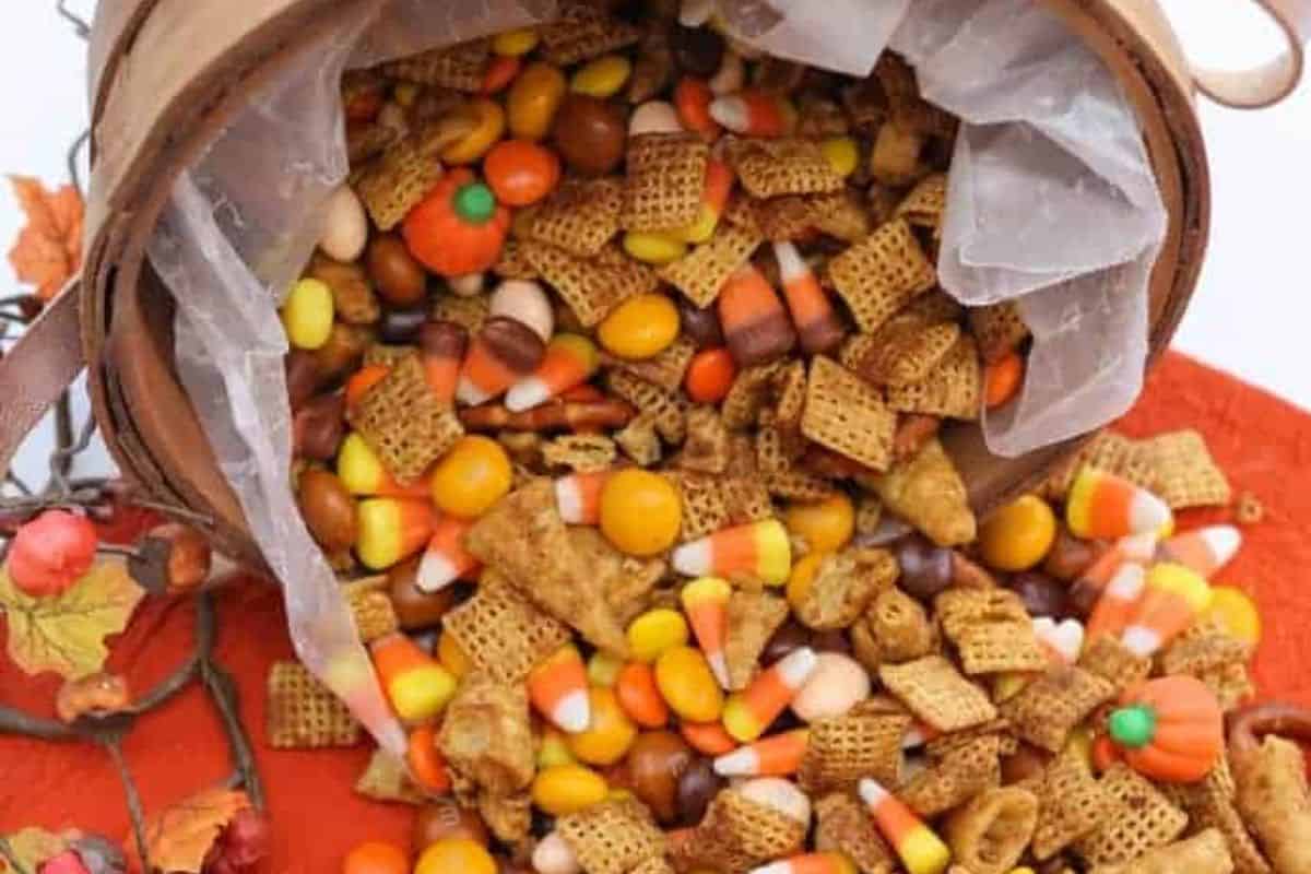 A basket tipped over, spilling a mix of Chex cereal, candy corn, candy pumpkins, and chocolate candies onto a red surface—perfect inspiration for irresistible pumpkin spice recipes.