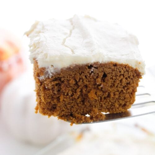 A square piece of pumpkin spice cake with white frosting is held up on a metal spatula, with a blurred background.