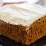 A slice of moist pumpkin spice cake with white frosting on top, served on a black plate.