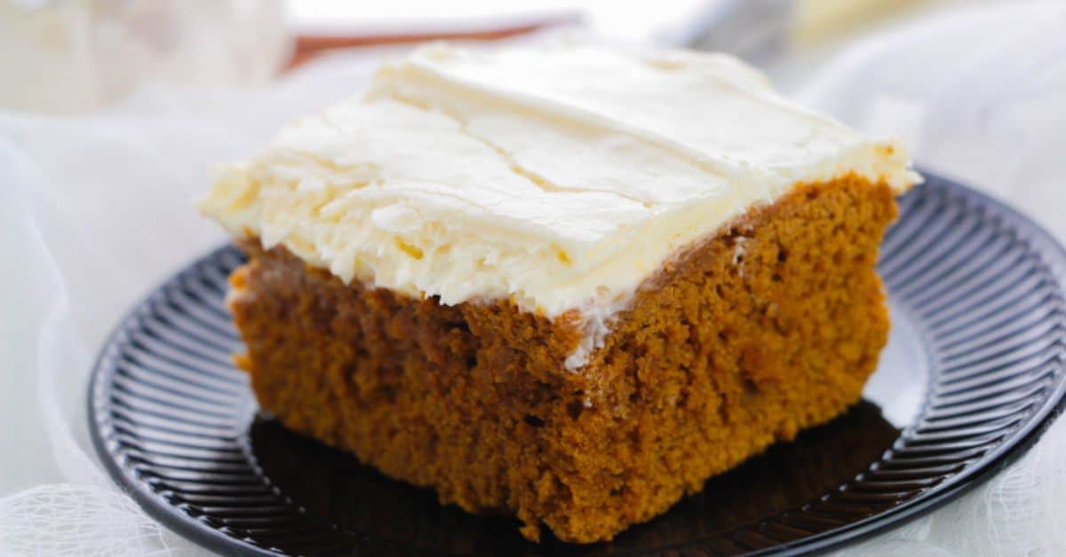 A slice of moist pumpkin spice cake with white frosting on top, served on a black plate.