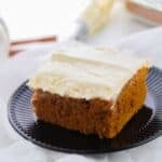 A single square piece of dessert topped with luscious frosting, sits on a black plate with white fabric underneath and baking tools in the background.