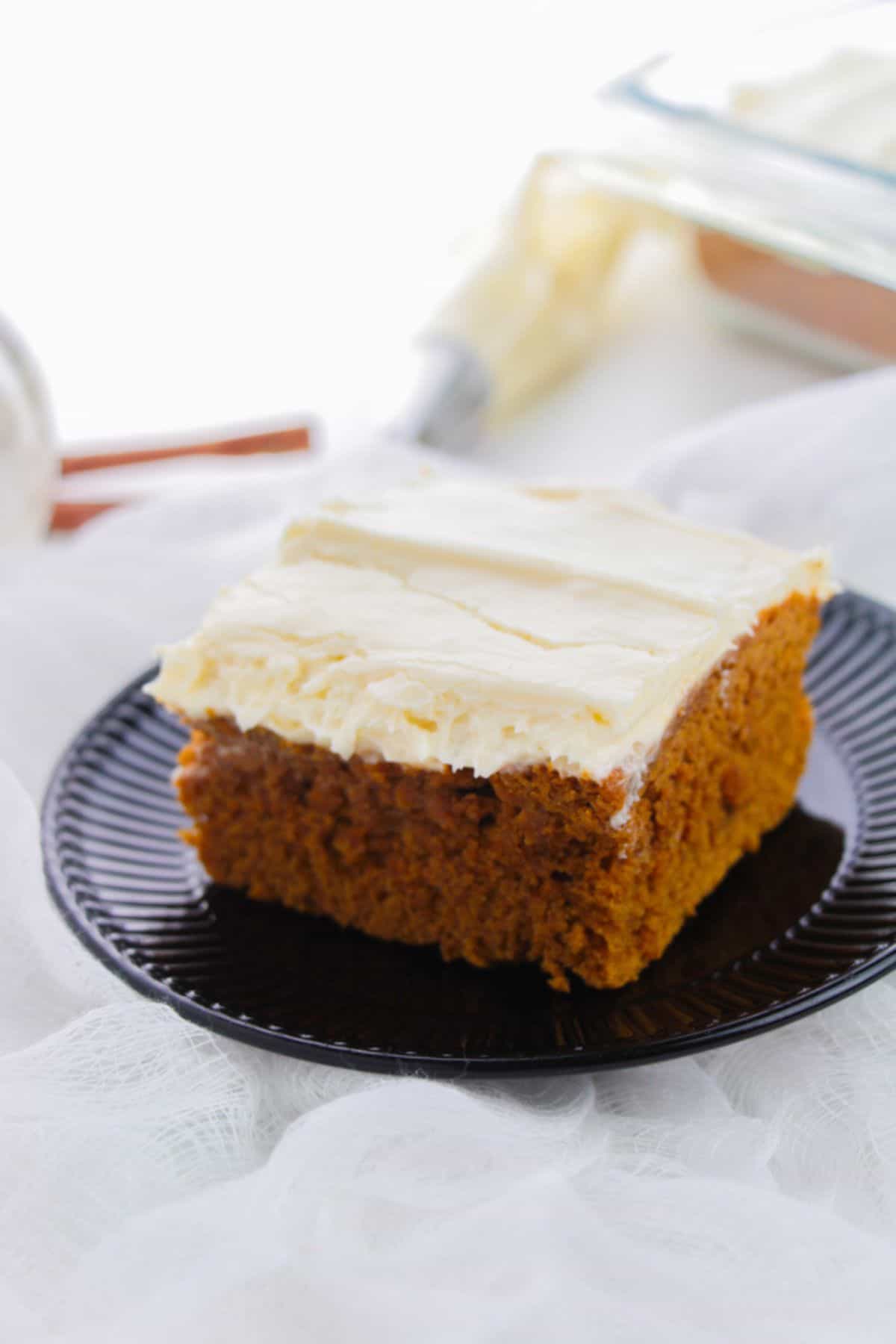 A single square piece of dessert topped with luscious frosting, sits on a black plate with white fabric underneath and baking tools in the background.
