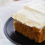 A slice of frosted pumpkin spice cake sits on a black plate with a white fabric background.