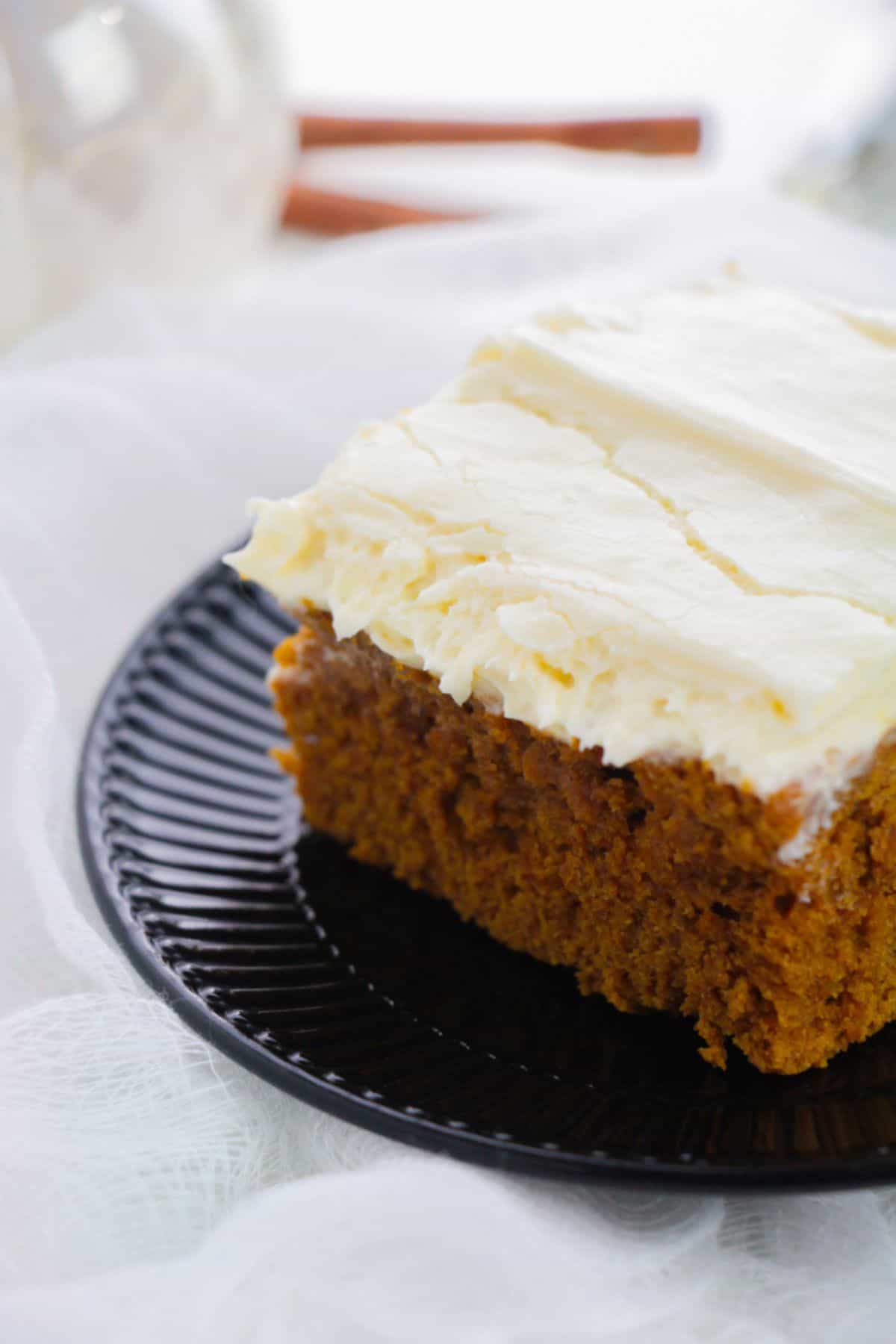 A slice of frosted pumpkin spice cake sits on a black plate with a white fabric background.