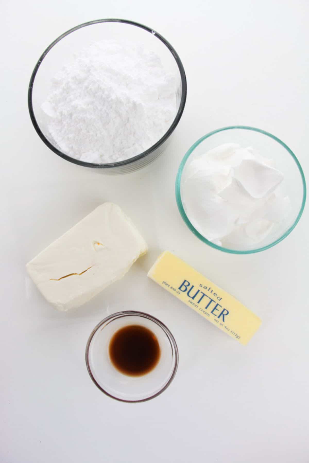 Five baking ingredients on a white surface: a bowl of powdered sugar, a bowl of shortening, a block of cream cheese, a stick of butter, and a small bowl of vanilla extract—perfect for frosting your favorite pumpkin spice cake.