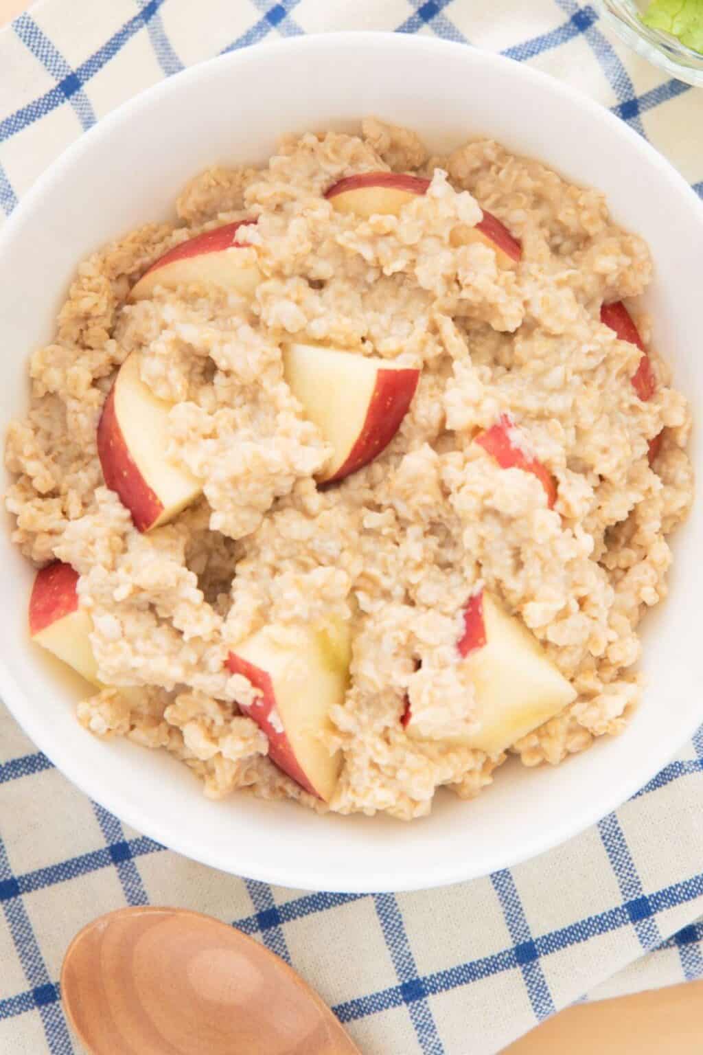 A white bowl filled with apple cinnamon oatmeal and fresh apple slices sits on a blue and white checkered cloth, with a wooden spoon nearby.