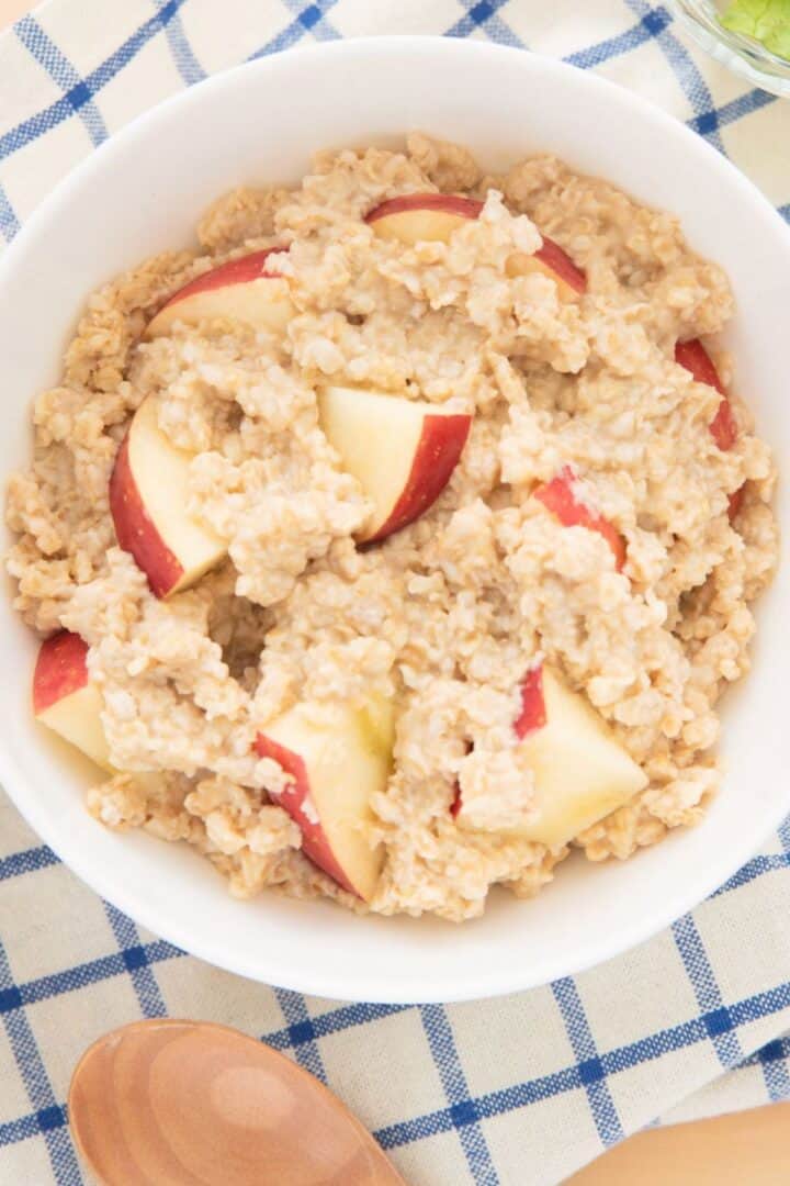 A white bowl filled with apple cinnamon oatmeal and fresh apple slices sits on a blue and white checkered cloth, with a wooden spoon nearby.