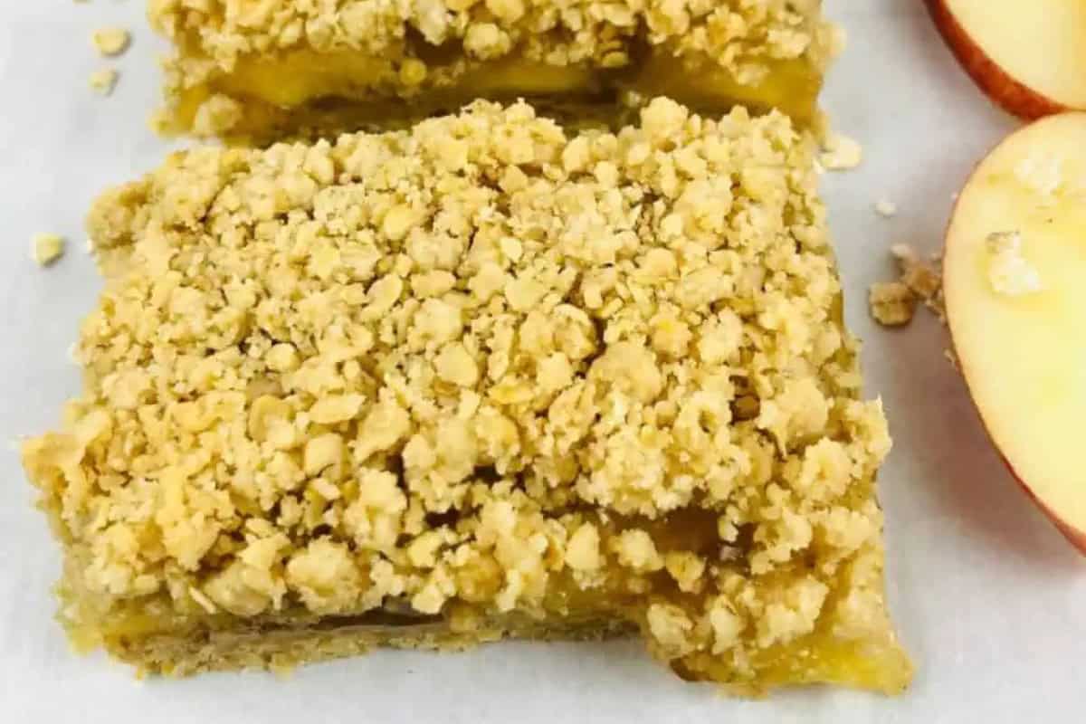 A close-up of two apple crumble bars with a crumbly oat topping next to a sliced apple on a white surface.