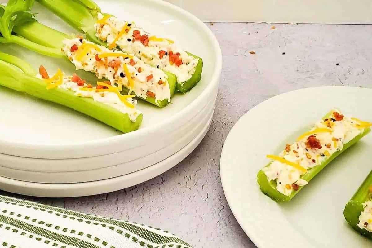 Celery sticks filled with cream cheese and topped with shredded cheese and bacon bits are arranged on white plates. There is 4 plates stacked on top of each other on the left side with 3 celery sticks with some leaves and stalks on top, while 2 celery sticks without stalks or leaves can be seen on the right side resting on a single plate.