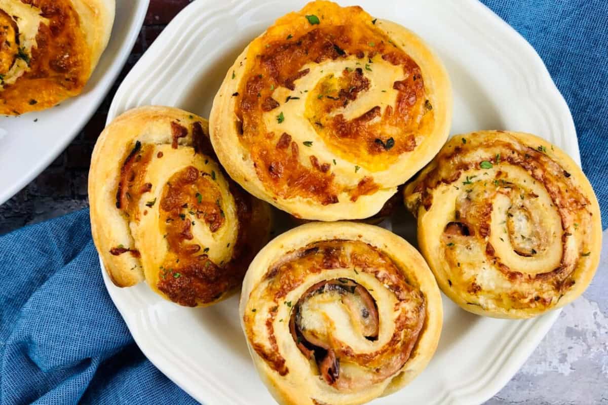 A white plate with four golden-brown pinwheel pastries filled with ham, cheese and herbs, placed on a blue cloth napkin.