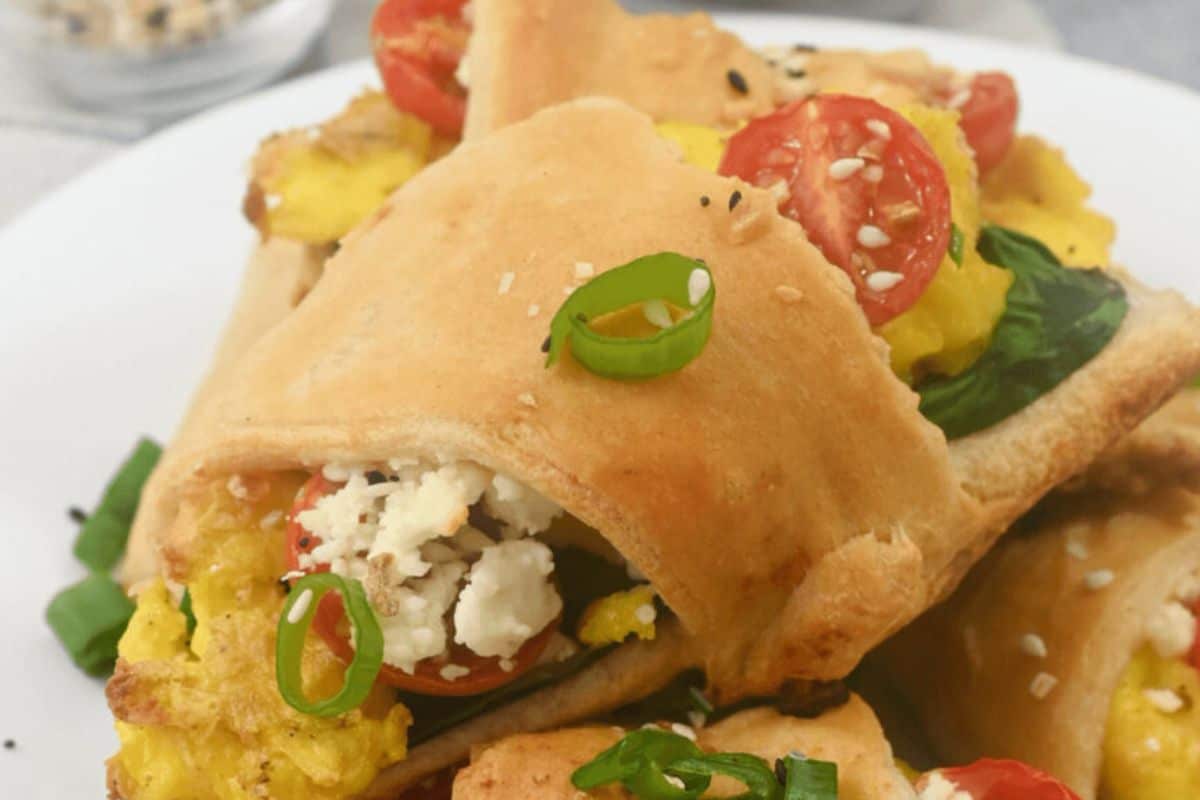 A perfect plate of breakfast croissant rollups filled with scrambled eggs, cherry tomatoes, spinach, and crumbled cheese, garnished with sesame seeds and sliced green onions.
