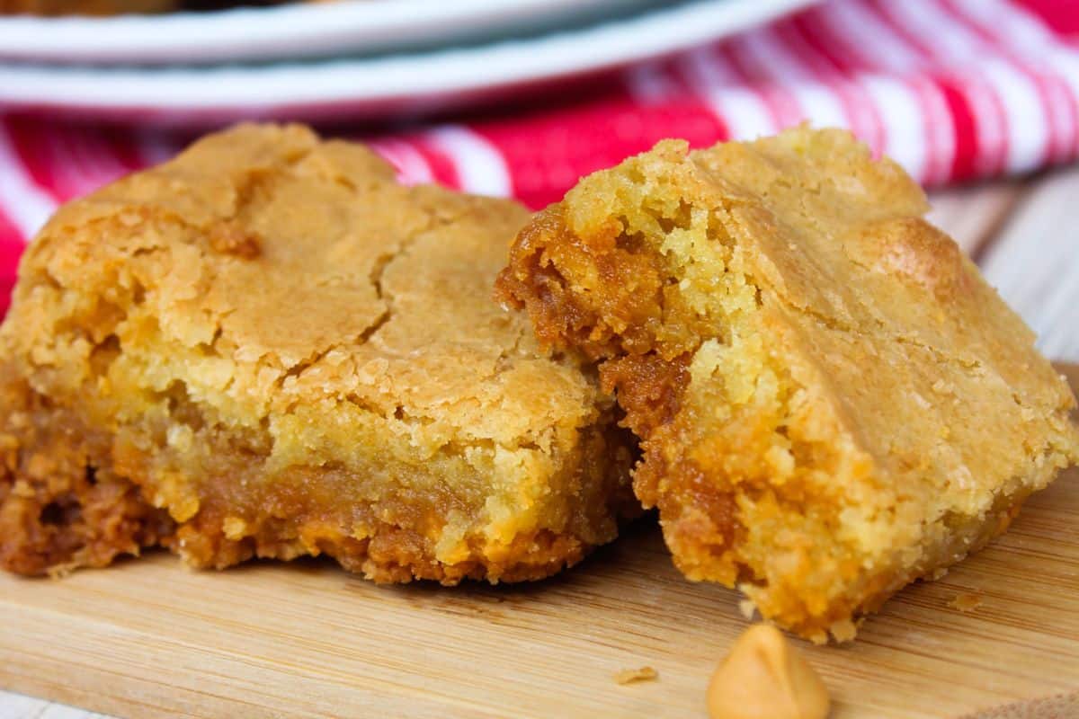 Two close-up squares of peanut butter blondies with a crumbly, golden exterior, placed on a wooden surface. A red and white striped cloth is in the background, evoking the charm of old-fashioned dessert recipes.