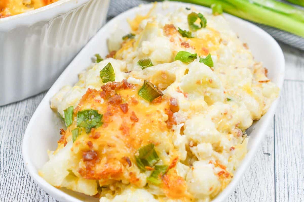 A dish of cheesy baked cauliflower garnished with green onions in a white rectangular bowl on a wooden surface, reminiscent of grandma recipes from classic vintage casserole dishes.